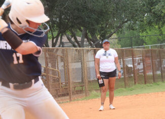 New College of Florida softball coach Cheyenne Nunez watches an at-bat during the team’s game against the University of Fort Lauderdale. The batter is closer to the camera and out of focus. Nunez is standing further away in a white T-shirt, black shorts and a visor. She is in focus.