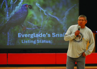 Brighton Community Culture advisor Johnnie Jones talks to PECS students about the importance of respecting wildlife. Jones holds a microphone in his right hand in front of a projector showing a photo of a dark gray bird, the "Everglades Snail", with an orange beak and orange feet.