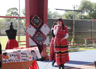 Durante Blais-Billie speaks at the MMIR prayer walk. She is wearing a red shirt and skirt and is holding a microphone.